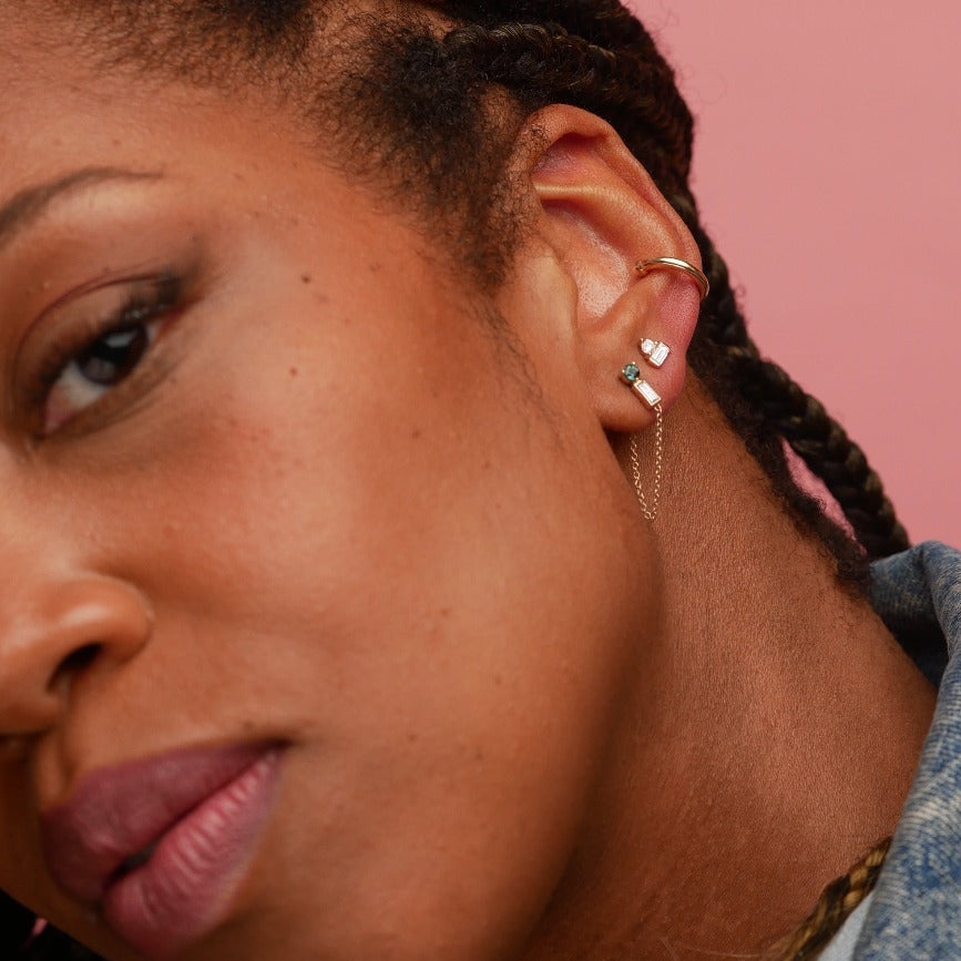 A close-up of a person's ear wearing a gold huggie hoop, a small gold diamond stud with a white heart, and a delicate gold chain earring, set against a solid pink background.